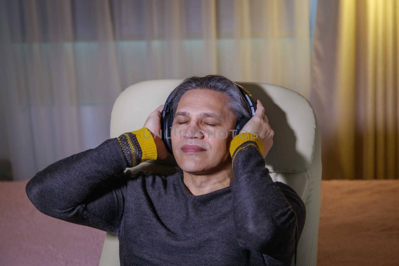 50-year-old man listens to music on headphones at home, sitting in a chair. Relaxing delight.