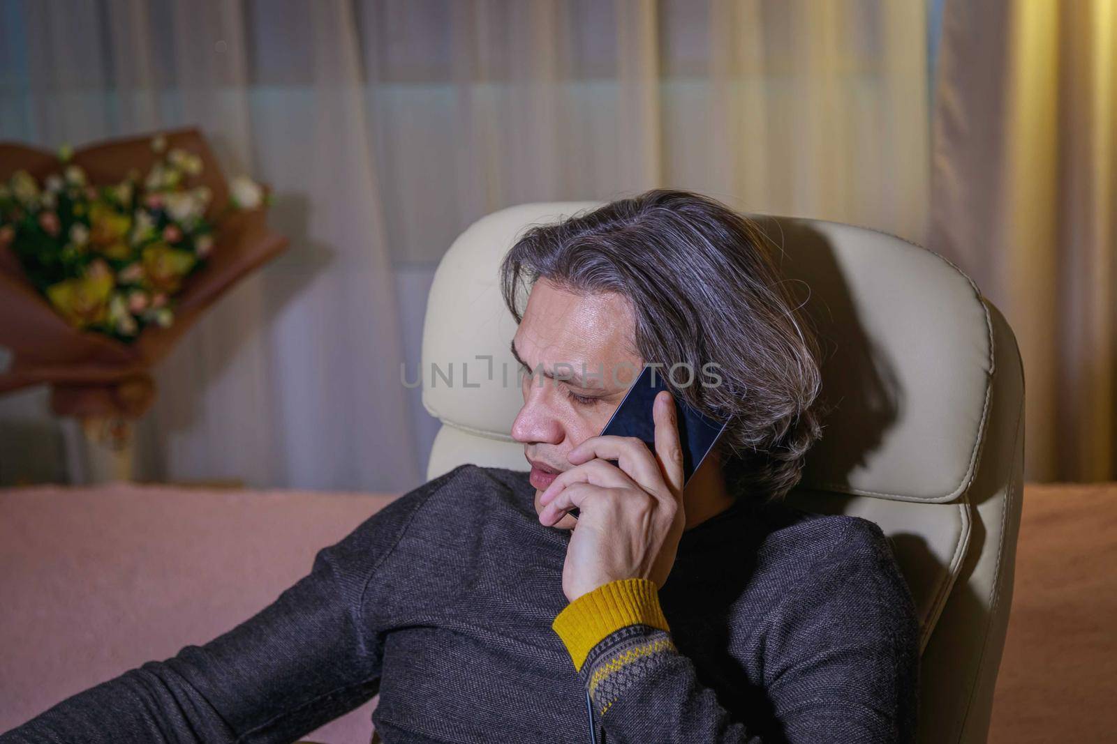 A 50-year-old man with long hair speaks on the phone at home, sitting in a chair. Relaxing delight.