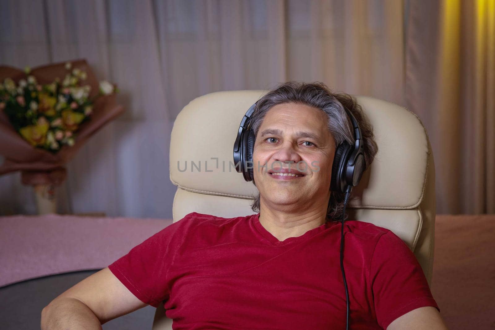 50-year-old man listens to music on headphones at home, sitting in a chair. Relaxing delight.