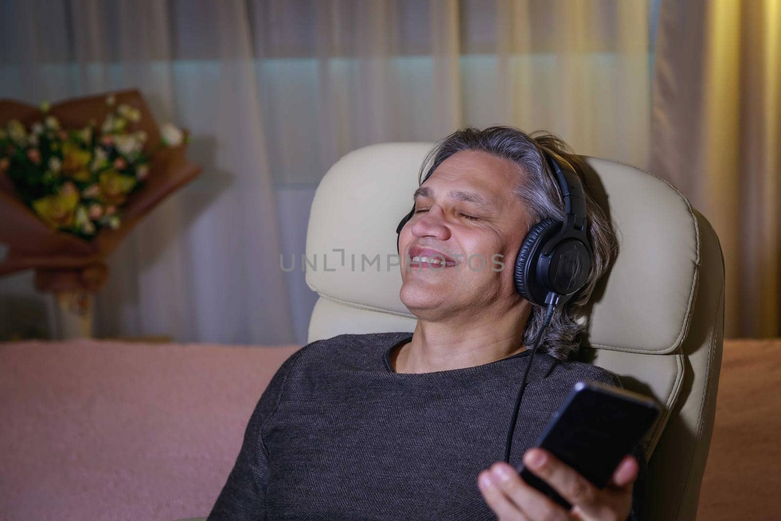 50-year-old man listens to music on headphones at home, sitting in a chair. Relaxing delight.