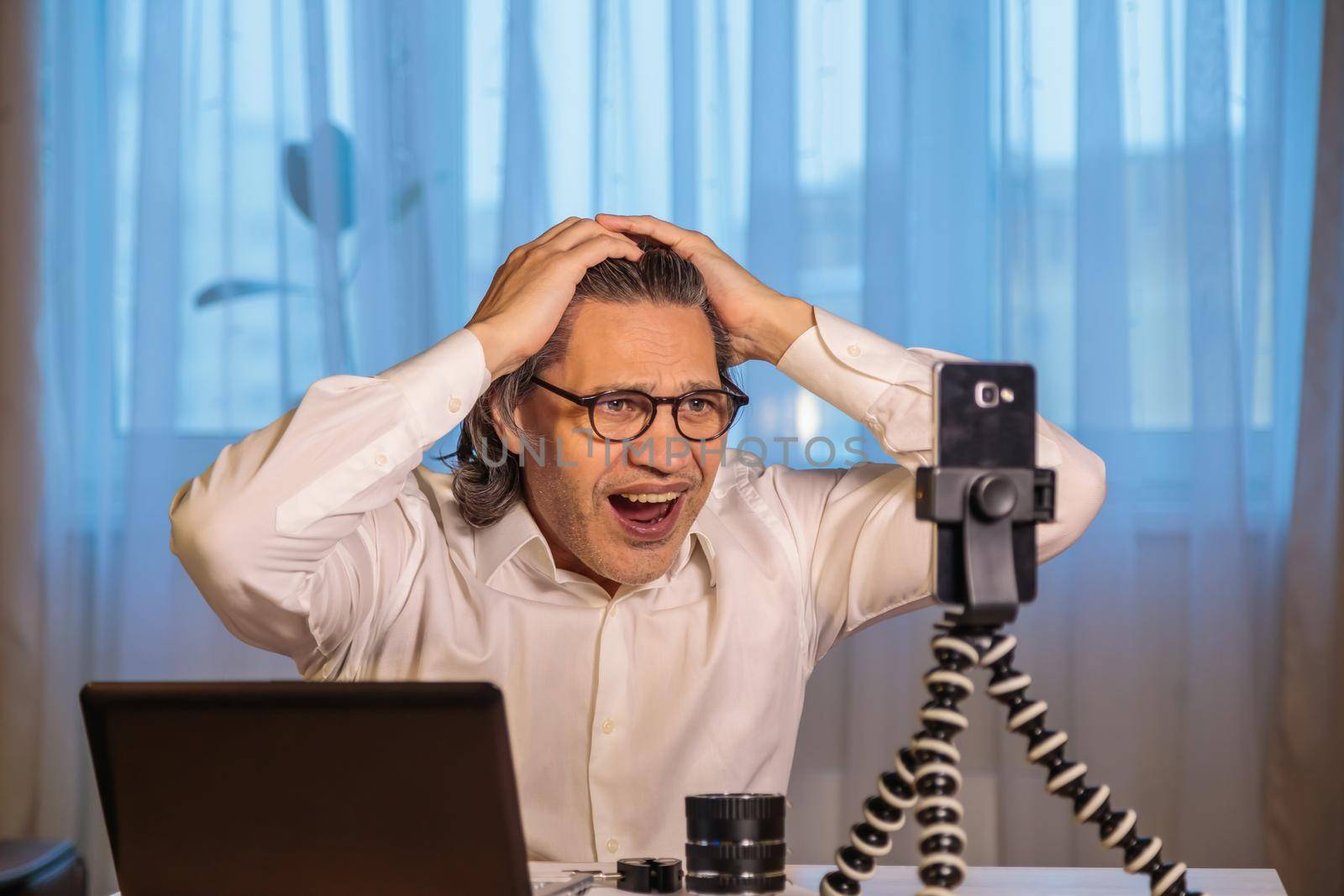 A 50-year-old blogger with glasses is recording an online seminar on his smartphone for his subscribers. With the help of modern devices, a computer and a smartphone, the master class is broadcast live.