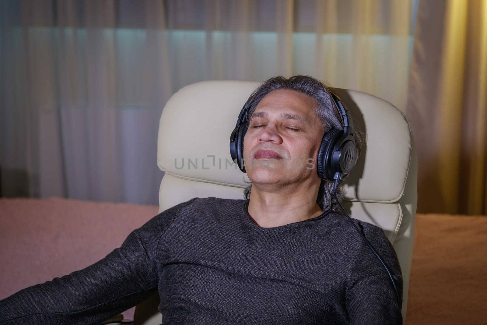 50-year-old man listens to music on headphones at home, sitting in a chair. Relaxing delight.