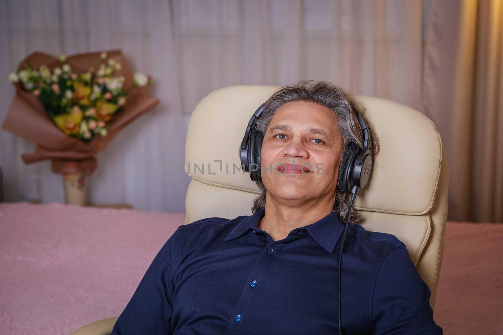 50-year-old man listens to music on headphones at home, sitting in a chair. Relaxing delight.