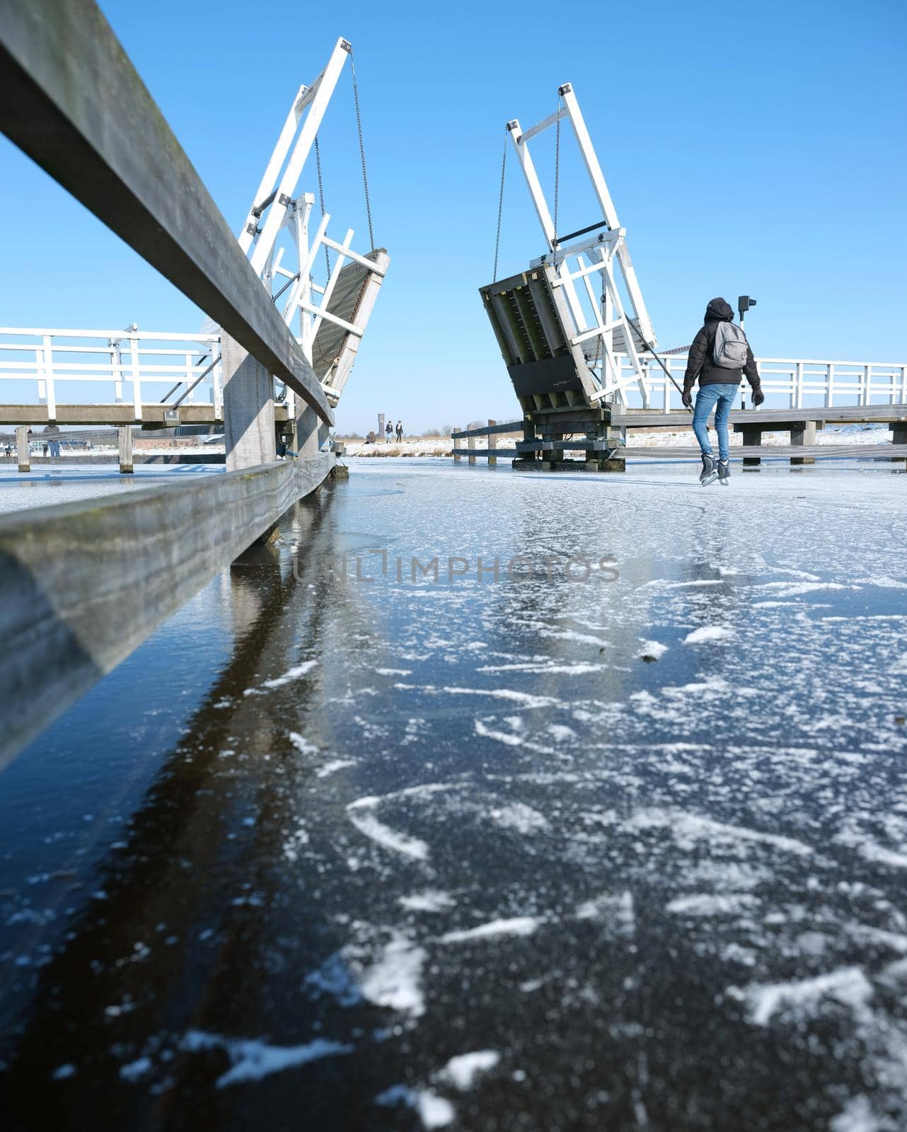 people skate through drawbridge in kinderdijk on frozen water of canal on sunny winter day in holland by ahavelaar