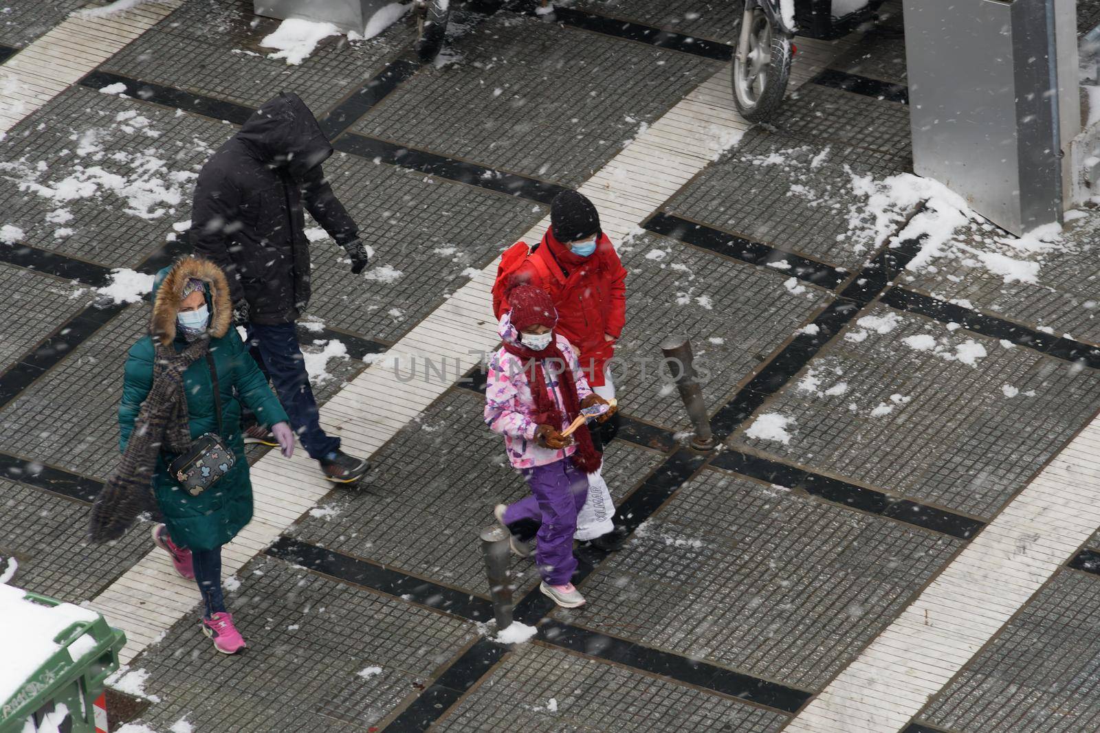 People in warm clothes and covid-19 masks walk under bad weather with snow falling.