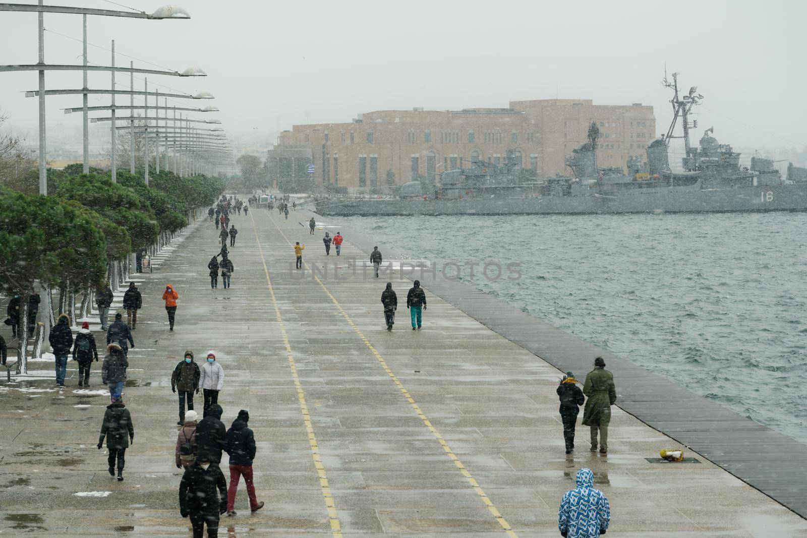 People in warm clothes and covid-19 masks walk at the waterfront with snow falling.