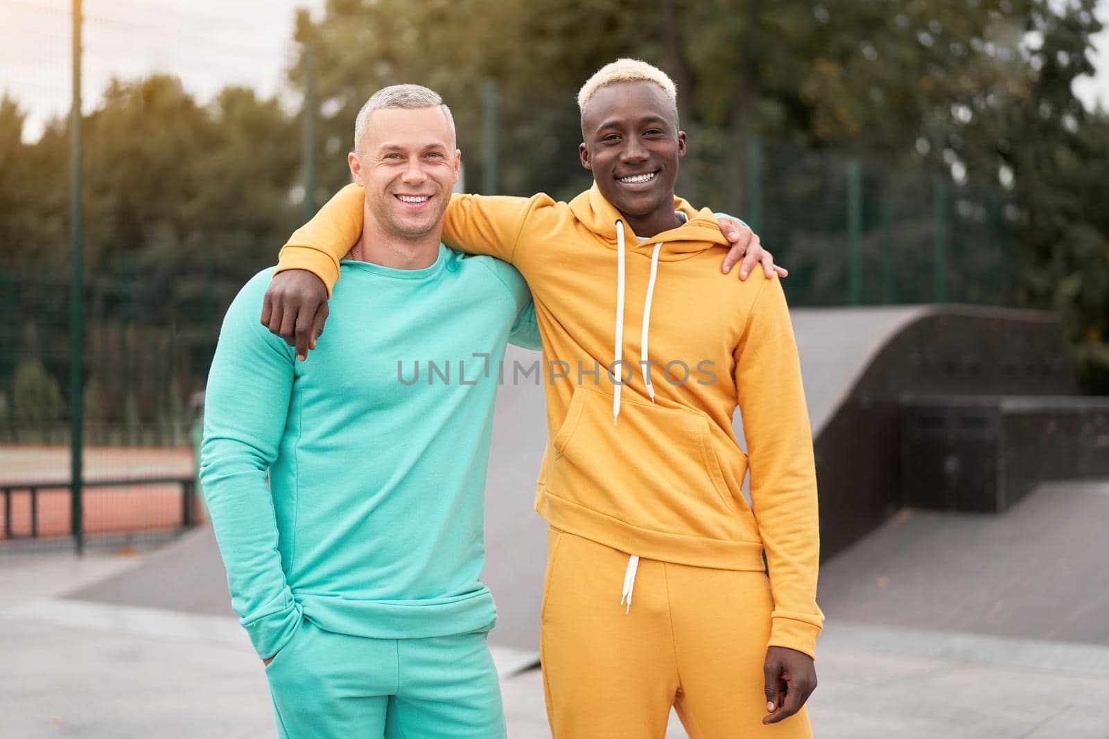 International friendship. Two happy diverse men in bright sportsuits standing outdoor, bonding together and smiling to camera in urban park