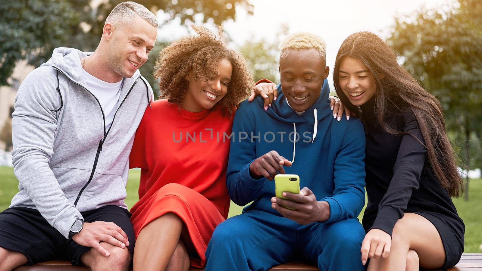 Young black guy sharing his mobile phone, showing funny joke in social media to his multiethnic friends, sitting together on bench in park
