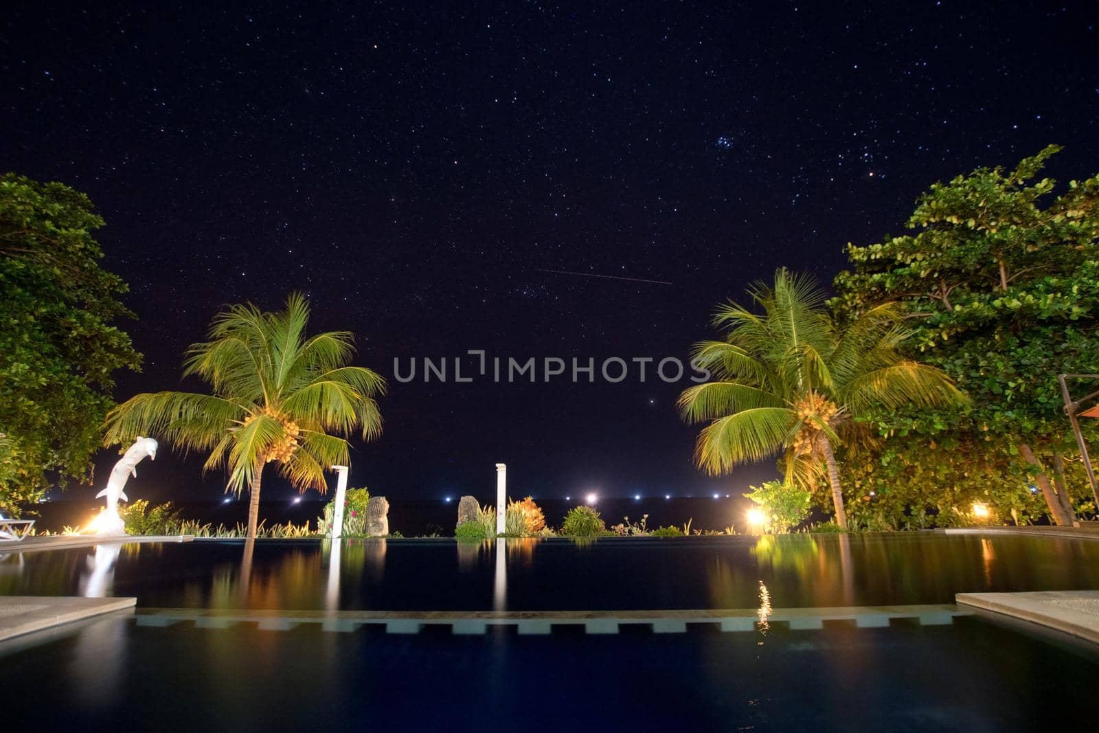 Luxury hotel with a swimming pool and palm trees under the starry sky. Long exposure, sky with stars, pool, sea, palm trees.
