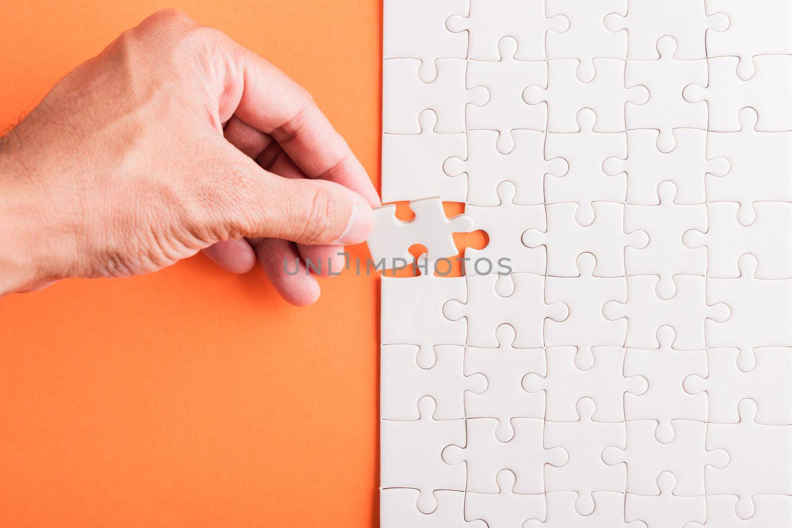 Top view flat lay of hand-holding last piece white paper jigsaw puzzle game last pieces put to place for solve problem complete mission, studio shot on an orange background, quiz calculation concept