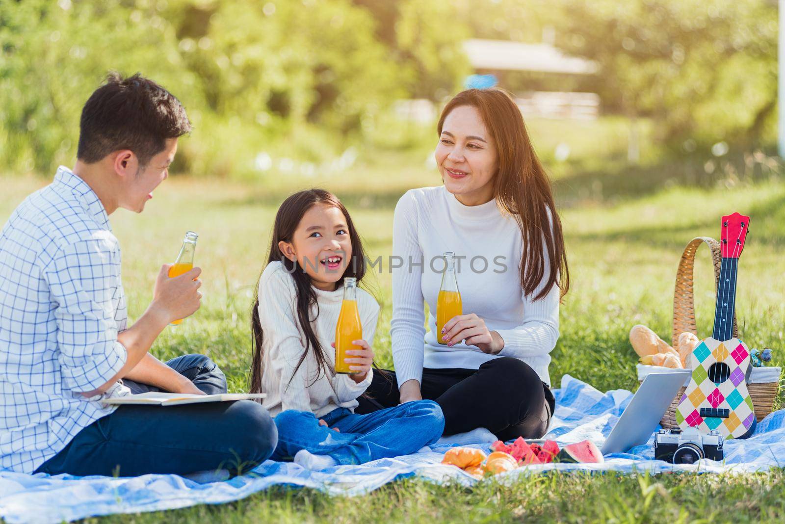 Happy family having fun outdoor sitting on picnic blanket drinking orange juice from glass bottle by Sorapop