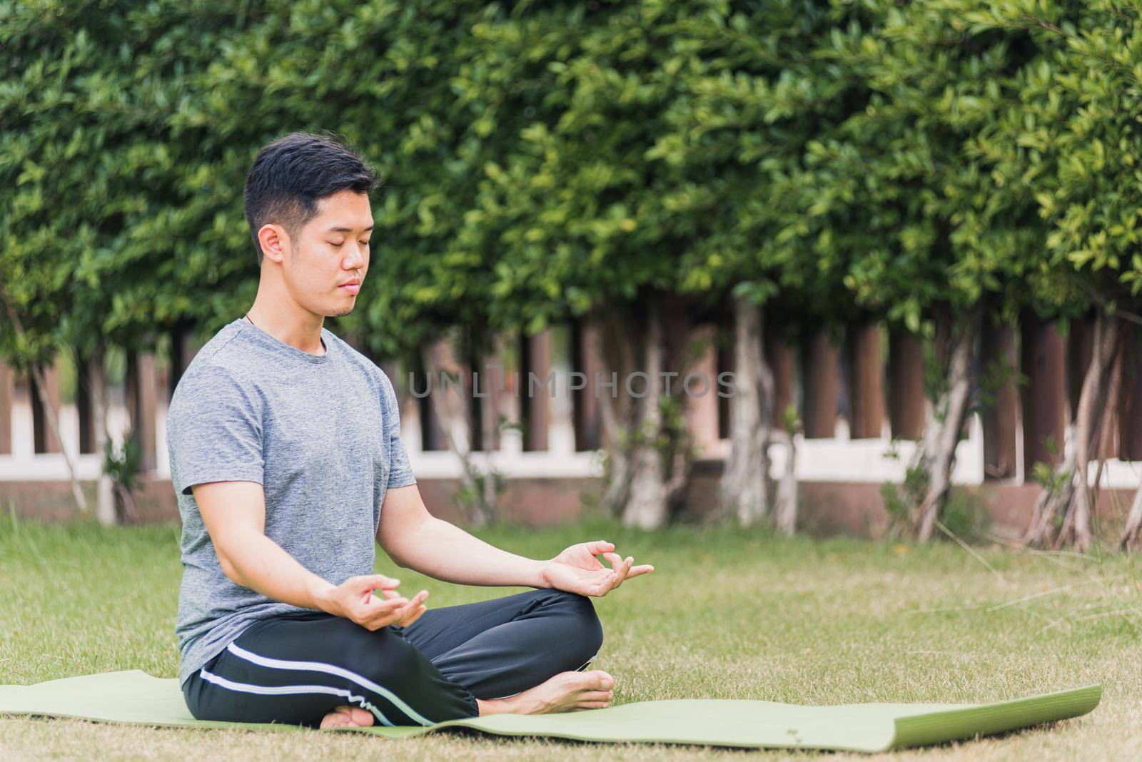 Asian young man doing yoga outdoors in meditate lotus pose sitting on green grass with closed eyes at the garden park, health care concept