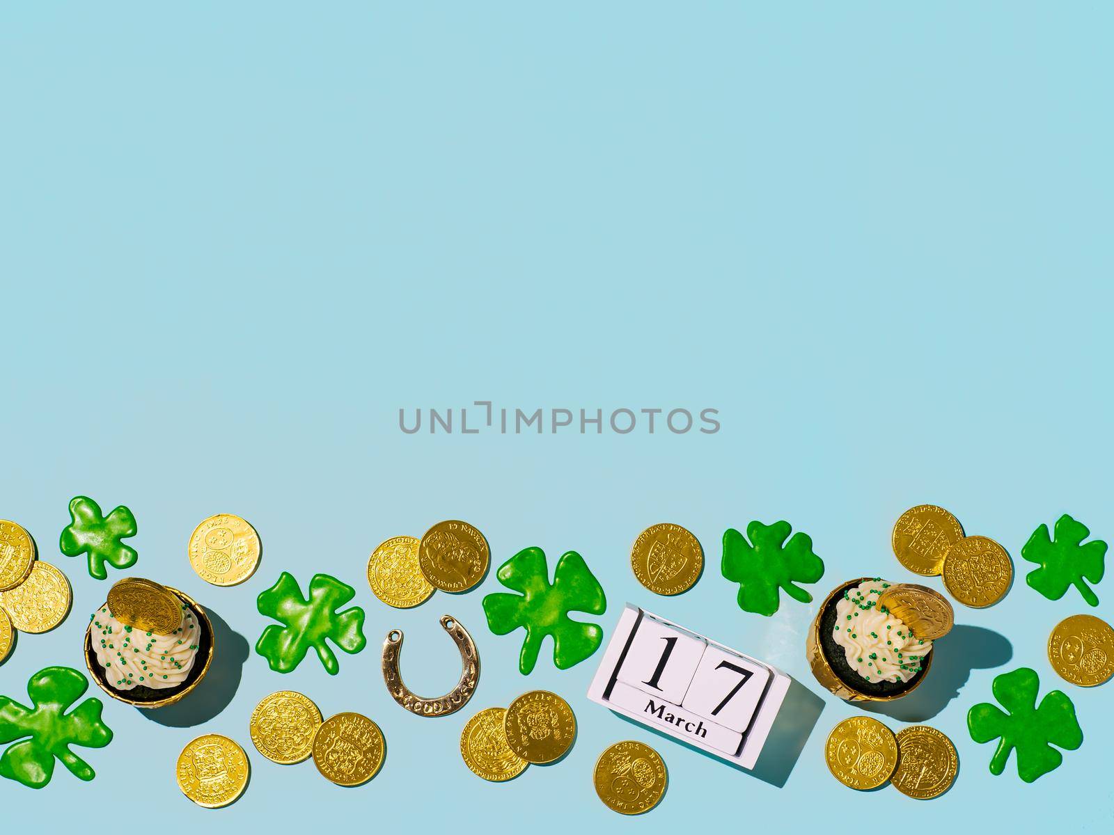 St Patrick's Day concept with copy space. Modern still life with lucky symbols and sweet food for Saint Patrick's Day party. Green velvet cupckake, shamrock, golden coins, horseshoe on blue background