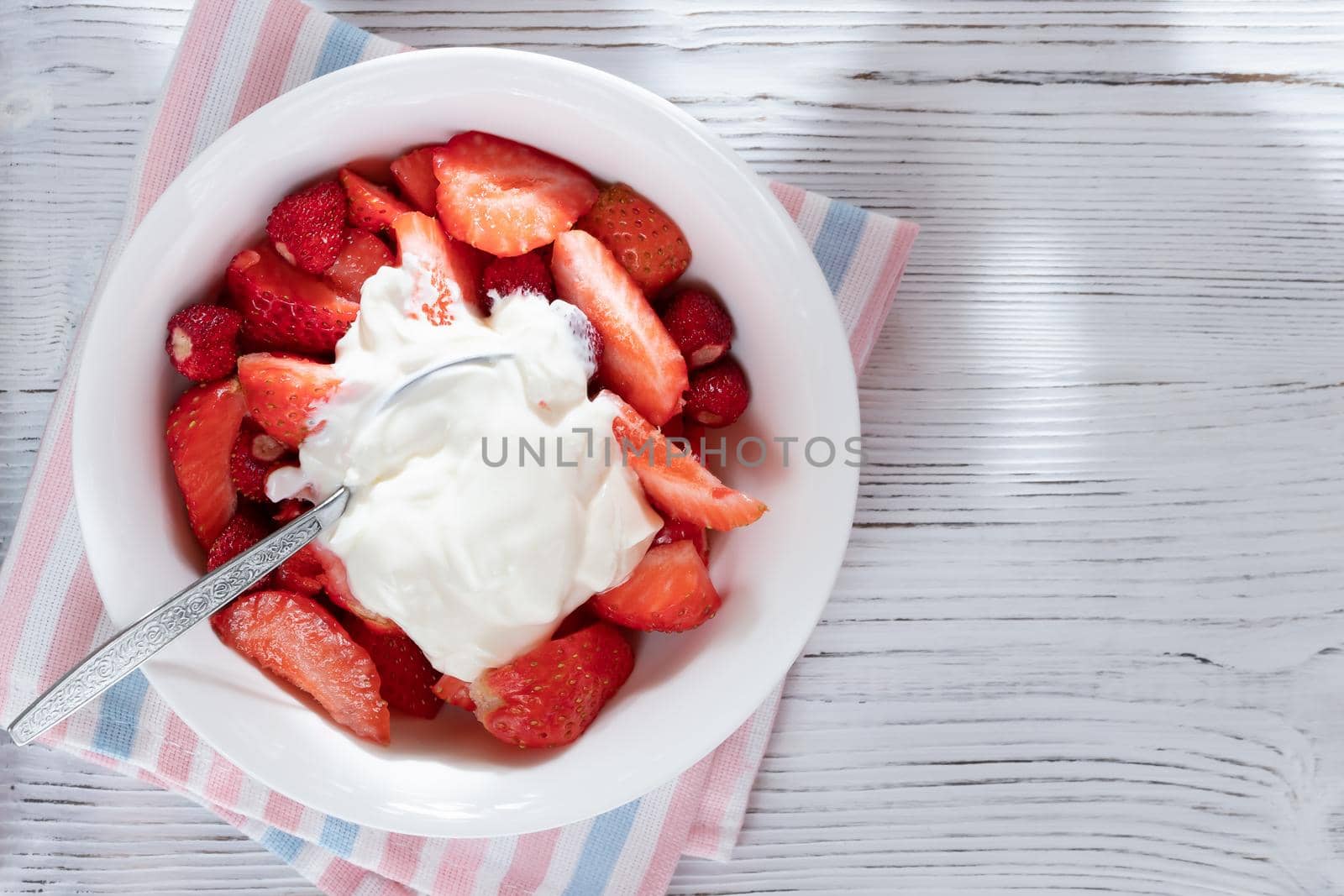 Natural sliced ripe strawberries in a white bowl with sour cream on a white wooden background. Top view, copy space.