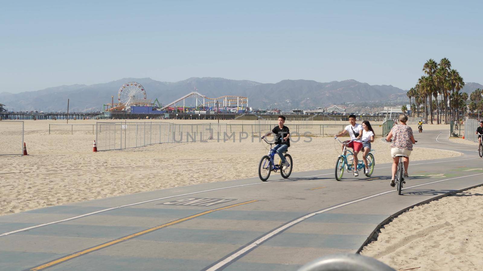 SANTA MONICA, LOS ANGELES CA USA - 28 OCT 2019: California summertime beach aesthetic, people walking and ride cycles on bicycle path. Amusement park on pier and palms. American pacific ocean resort.