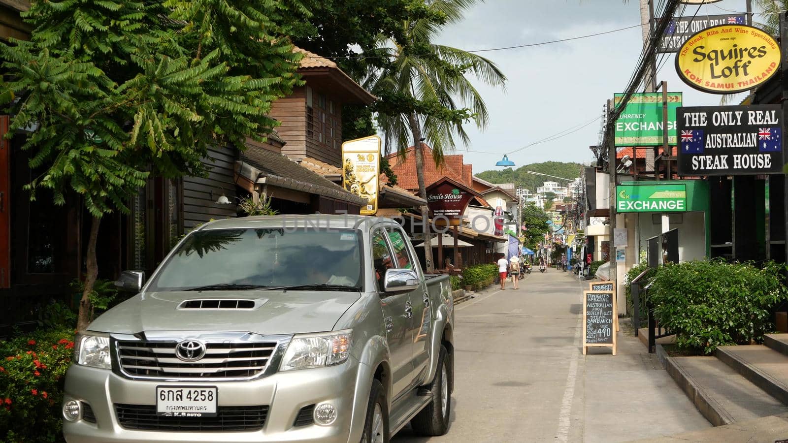 SAMUI ISLAND, THAILAND - MAY 27, 2019: Typical touristic street in Fisherman village with souvenir stores. View of calm lane of city in Asia with touristic shops in daytime. Thai people on motorbikes