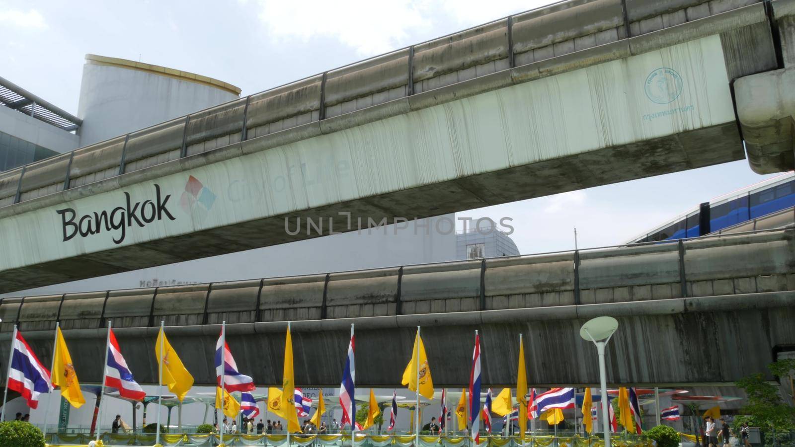 BANGKOK, THAILAND - 11 JULY, 2019: Pedestrians walking on the bridge near MBK and Siam Square under BTS train line. People in festive modern city decorated with waving national flags and royal symbol.