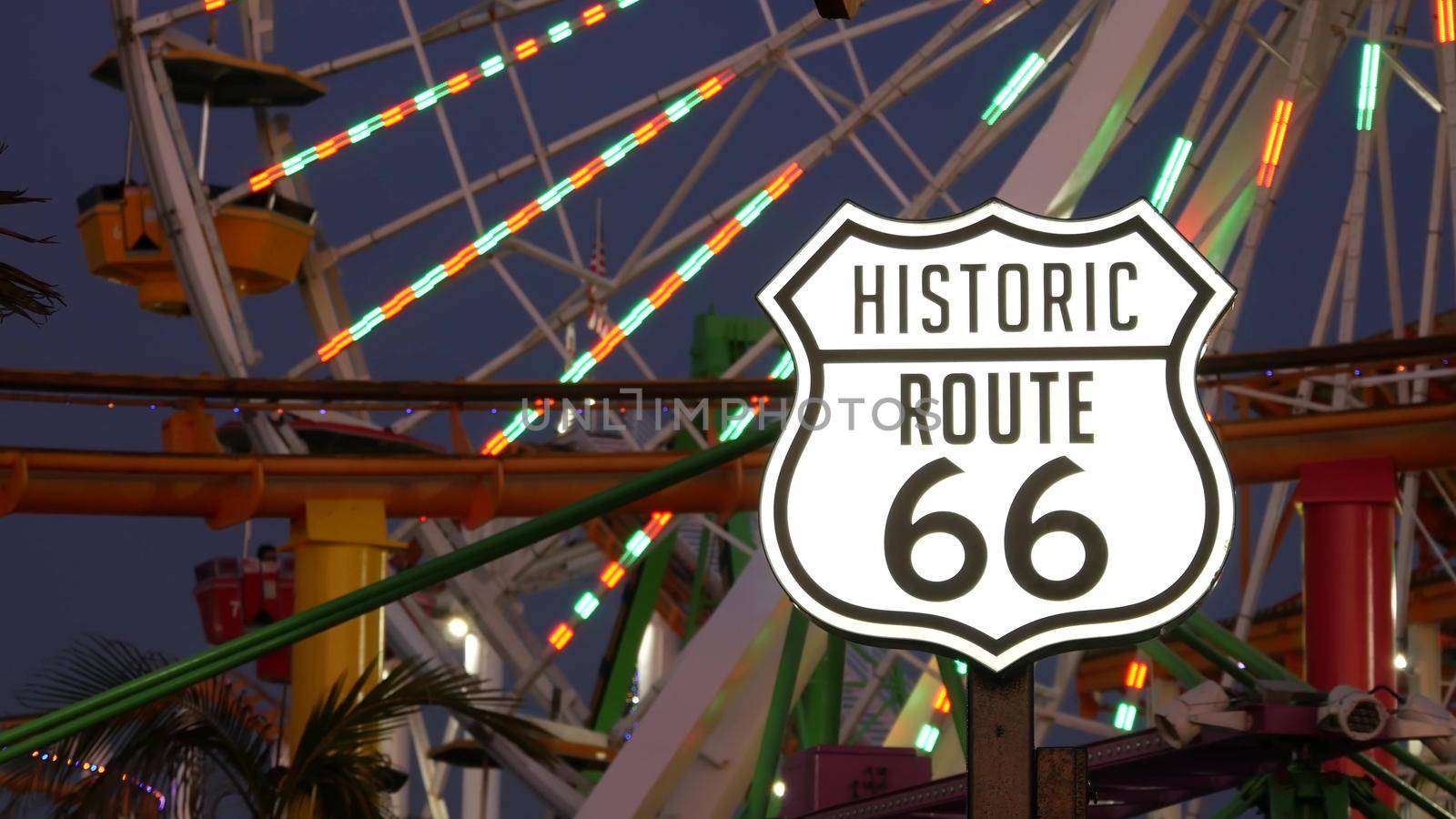 SANTA MONICA, LOS ANGELES, USA - 28 OCT 2019: Iconic road sign glowing, historic route 66. Famous california symbol, pier of pacific ocean resort. Illuminated festive ferris wheel in amusement park by DogoraSun