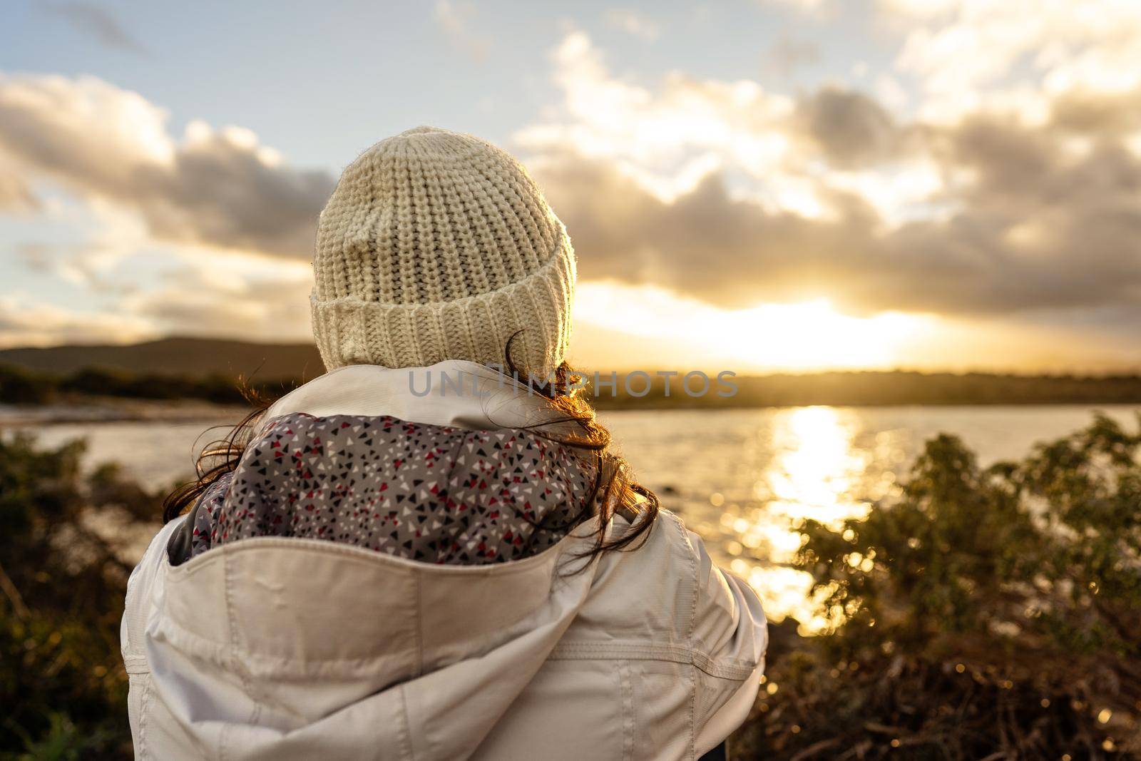 Pensive person view from back looking at the horizon. Unrecognizable woman seen from behind dressed in winter wool hooded clothes looks at a wonderful sunset or sunrise with clouds over the sea by robbyfontanesi