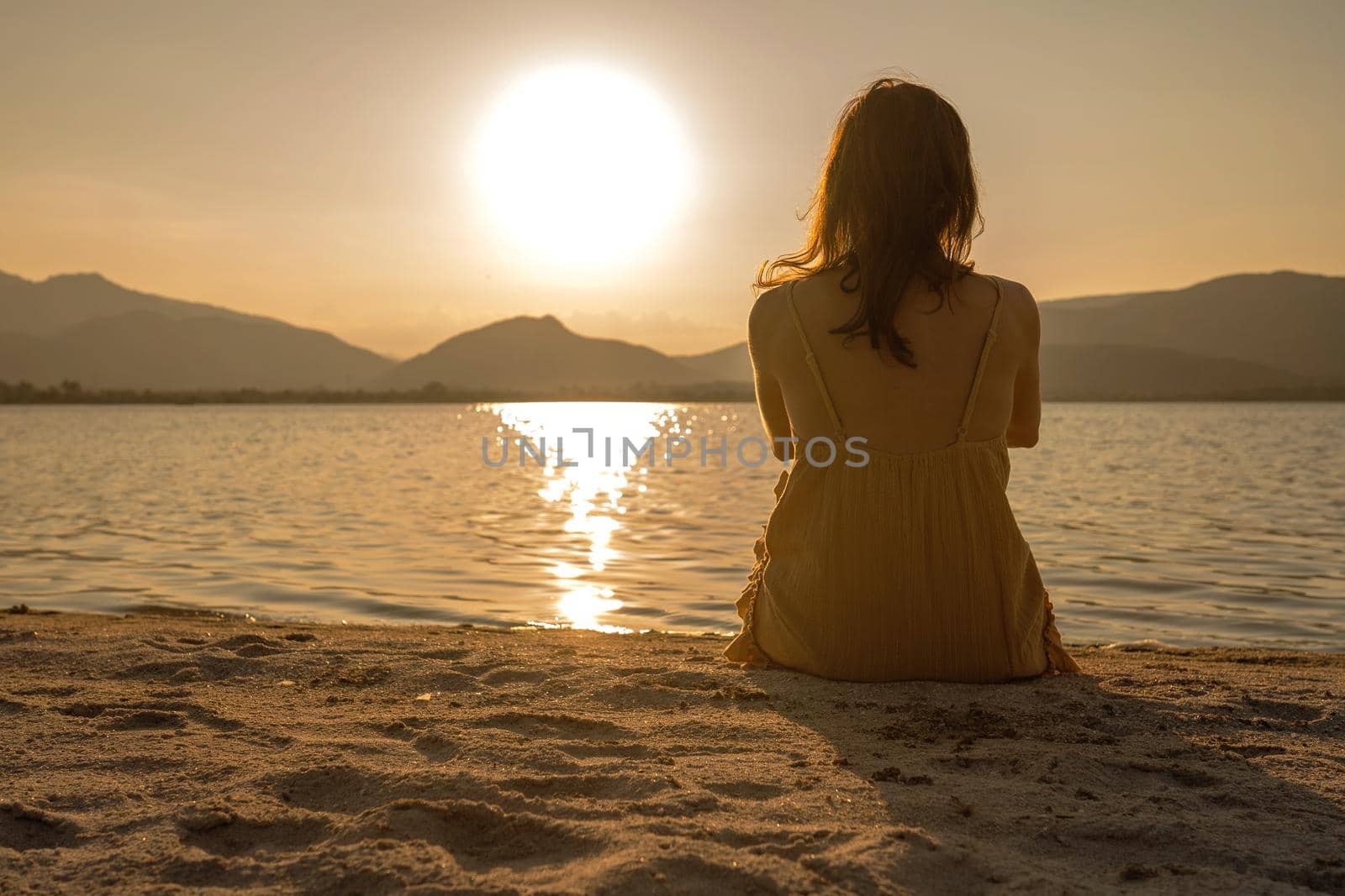 Unrecognizable lonely pensive woman sitting on the sand of seashore looking at the setting sun with light reflection in the water and orange sepia vintage photography effect. Dreaming person at dawn by robbyfontanesi
