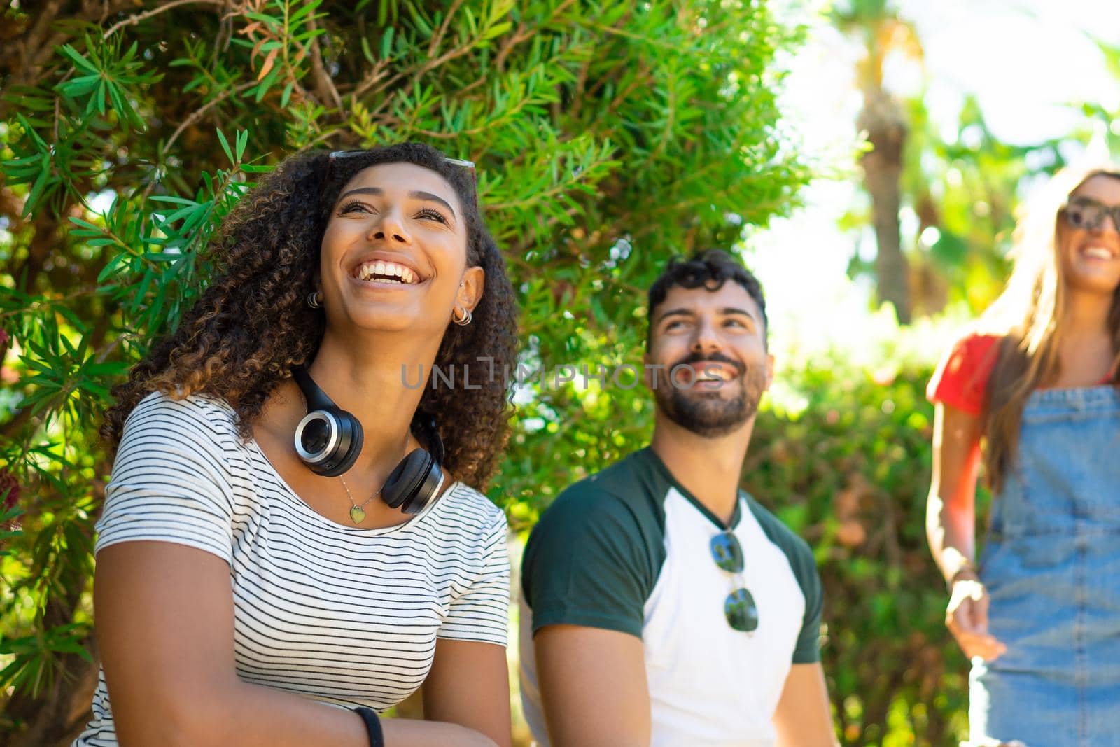 Afro american young woman with wireless headphones sitting in the city park with her millennials friends. Group of carefree hipster having fun in the summer sun and the nature in town