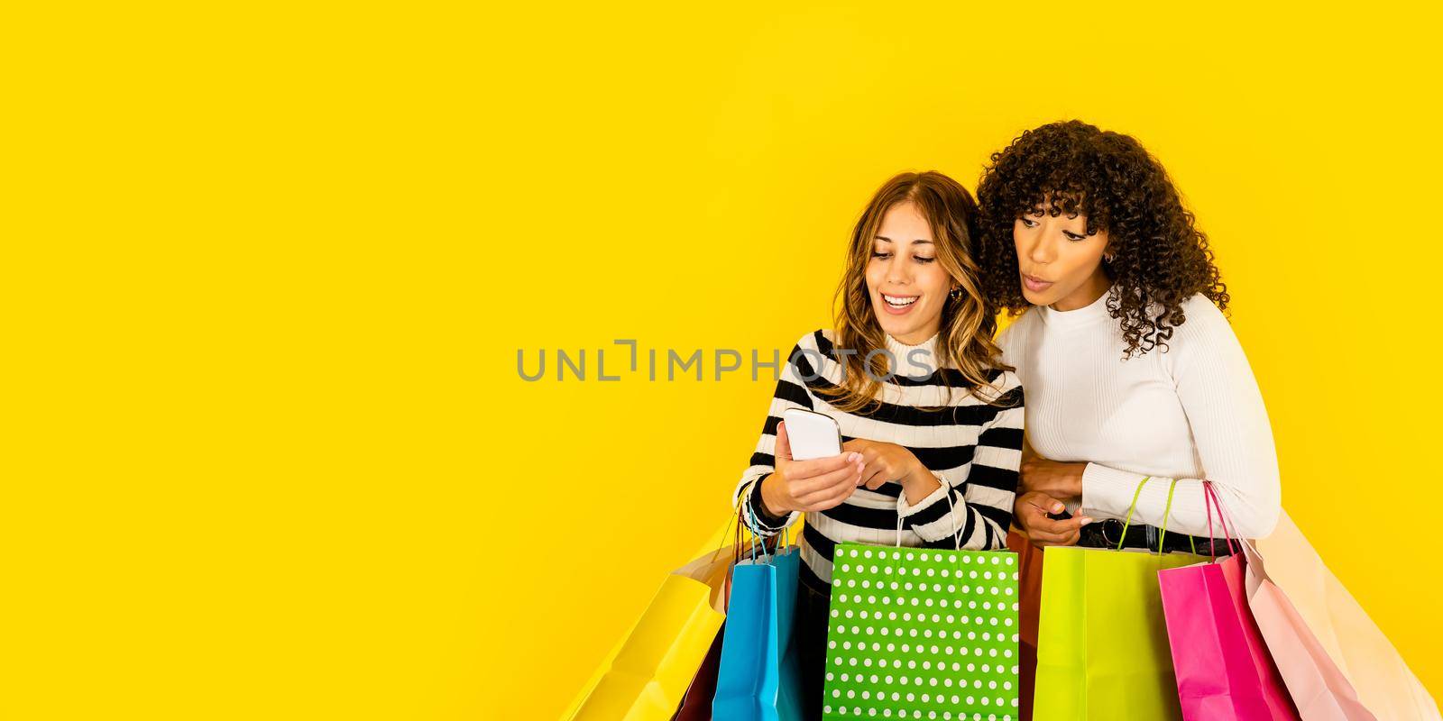 Afro american young surprised woman with wow face looking smartphone of her friend with several colored paper bags. Two shopaholic girls having fun with shopping online isolated on yellow copy space
