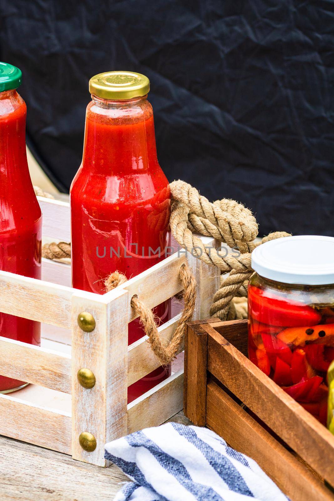 Glass jars with pickled red bell peppers and bottles with tomatoes sauce isolated in a rustic composition. Jars with variety of pickled vegetables preserved food concept.