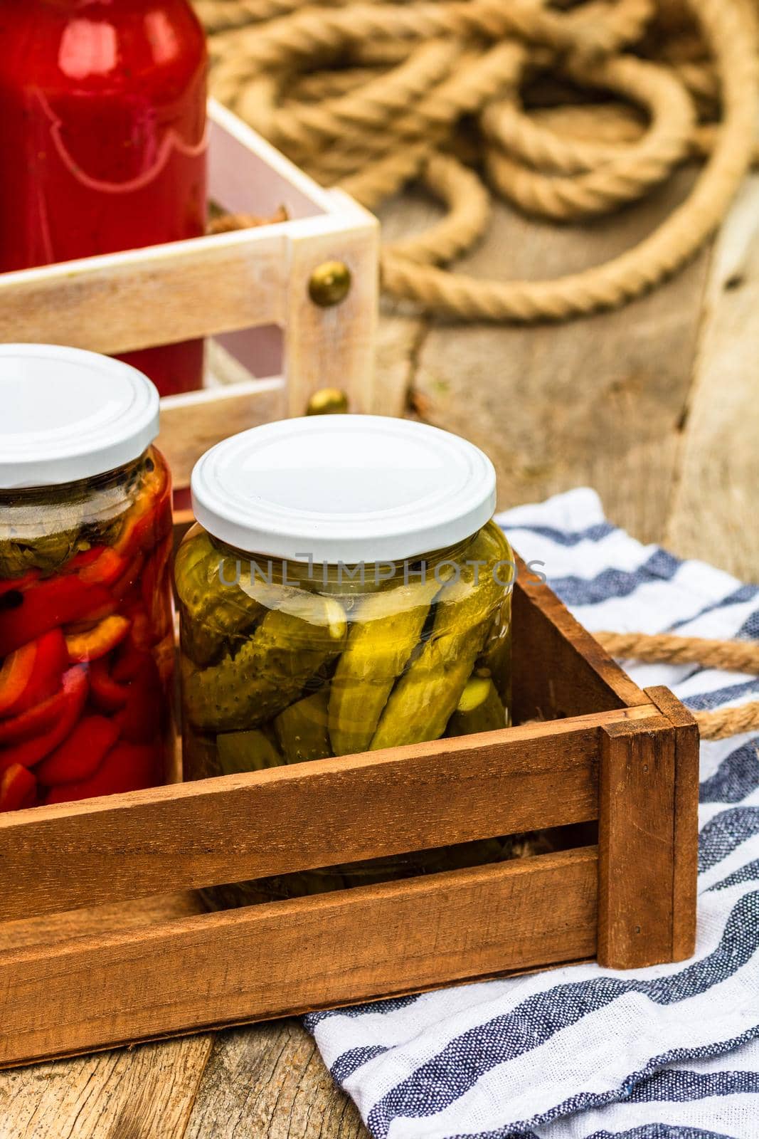 Wooden crate with glass jars with pickled red bell peppers and pickled cucumbers (pickles) isolated. Jars with variety of pickled vegetables. Preserved food concept in a rustic composition.
