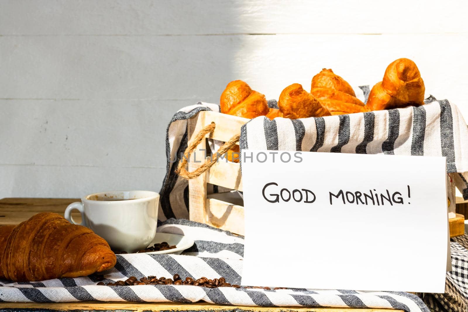 Coffee cup and buttered fresh French croissant on wooden crate. Food and breakfast concept. Morning message “good morning” on white board by vladispas