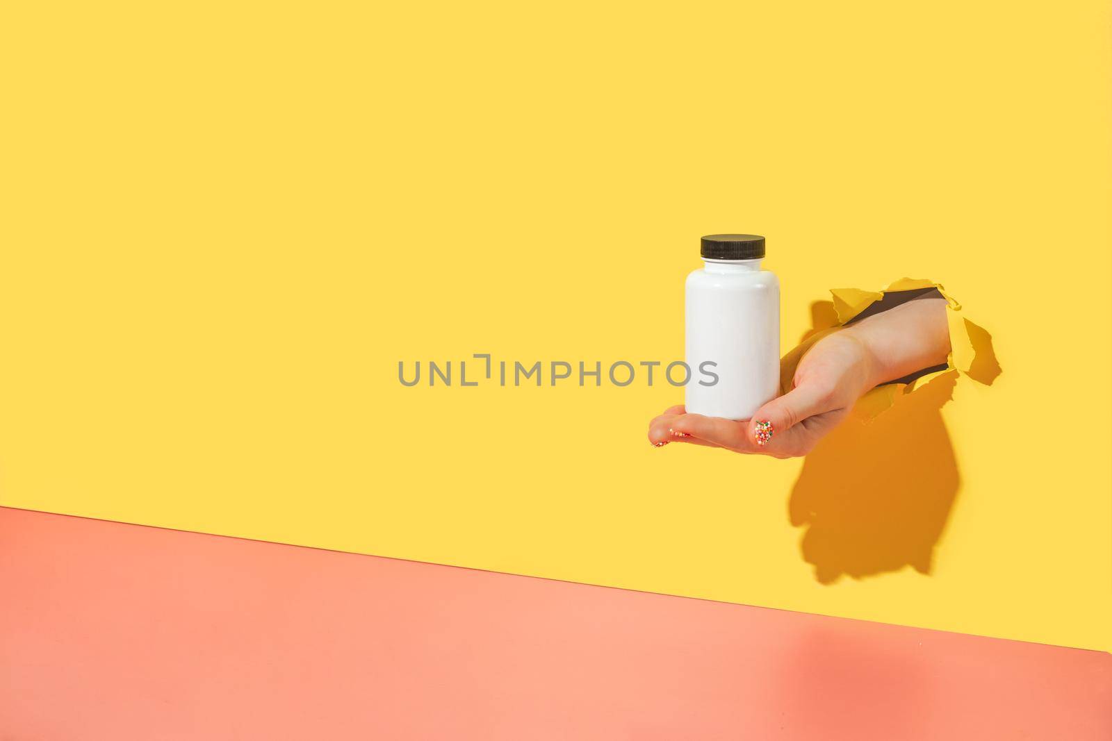 Empty white medical container for vitamins in female hand with sprinkle manicure through torn bright yellow paper background. Oral supplements, immune support, biohacking creative concept. Copy space