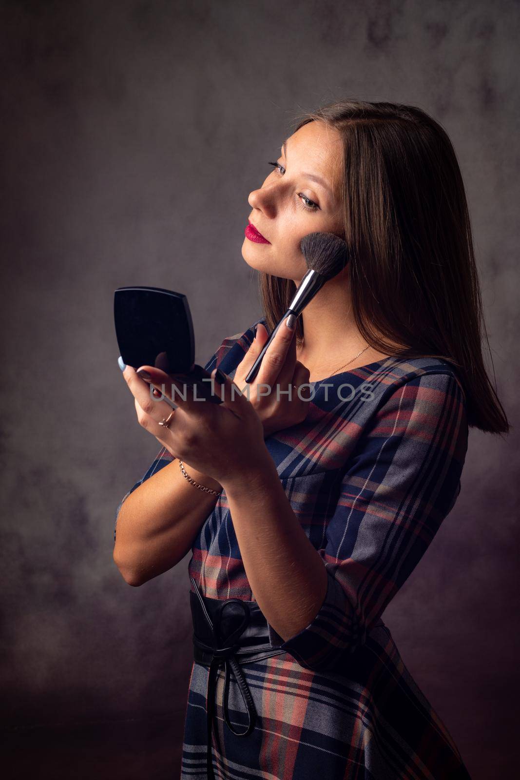 Girl powders her face with a brush looking in the mirror, studio photo on a gray background
