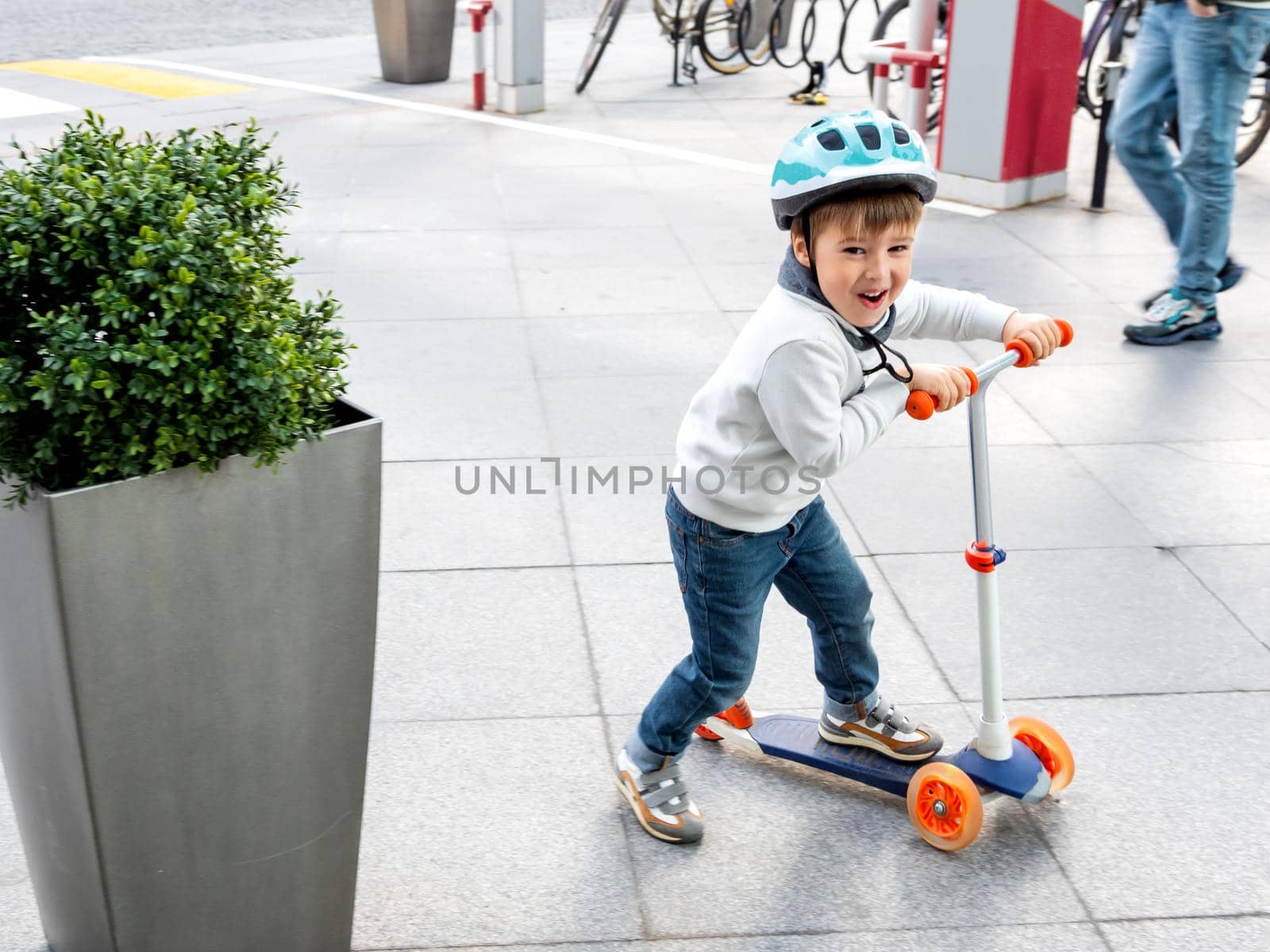 Toddler in helmet is riding scooter on parking lot with parked bicycles. Urban vehicle for active children. Leisure activity for boys and girls.