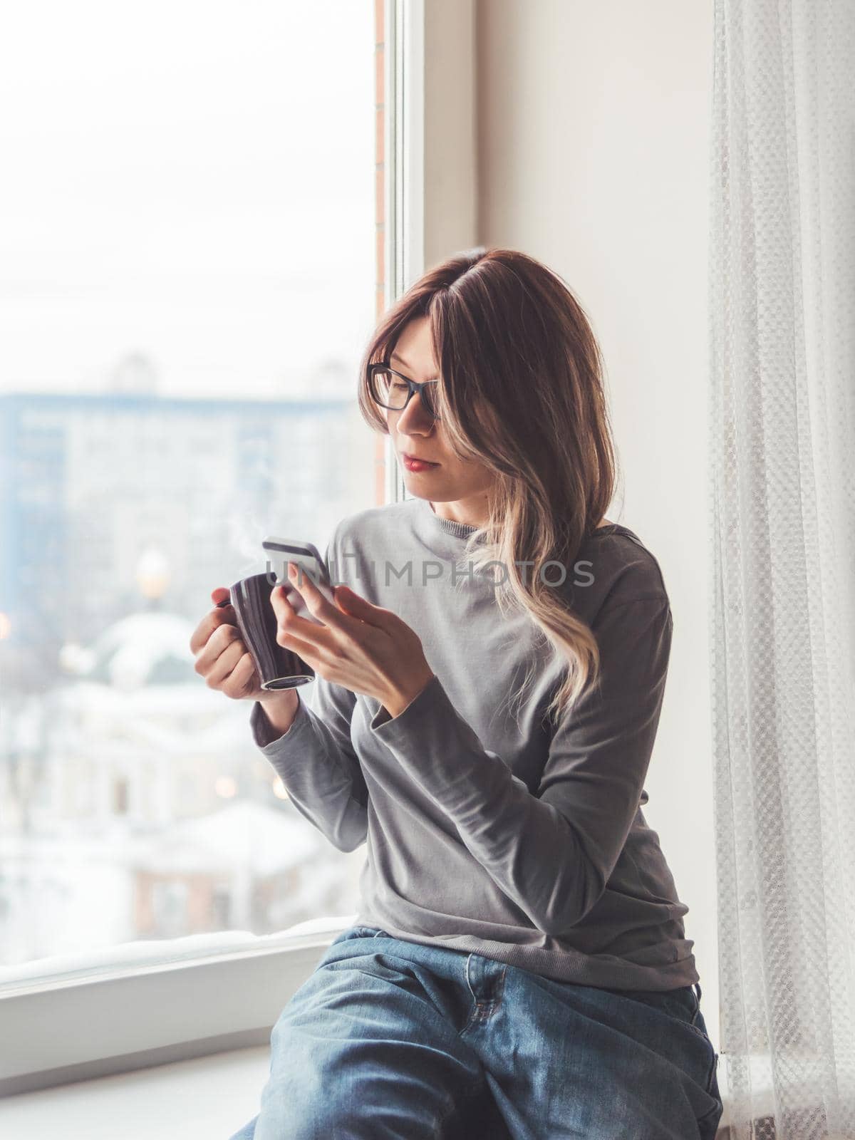 Thoughtful woman with eyeglasses and cup of hot coffee looks at her smartphone. Information in online media.