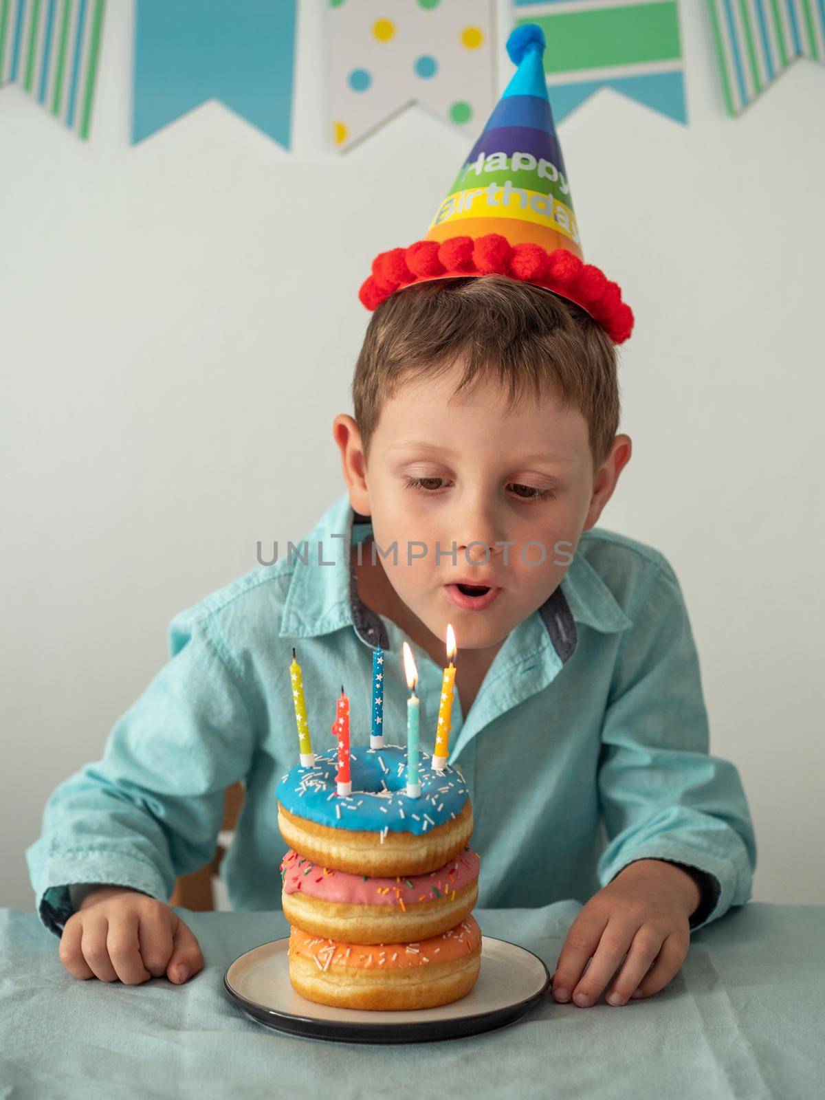 Five year old boy blows out the candles on festive donuts cake in his birthday. Happy little child and plate with doughnuts stack cake with candles on the table. Vertical.