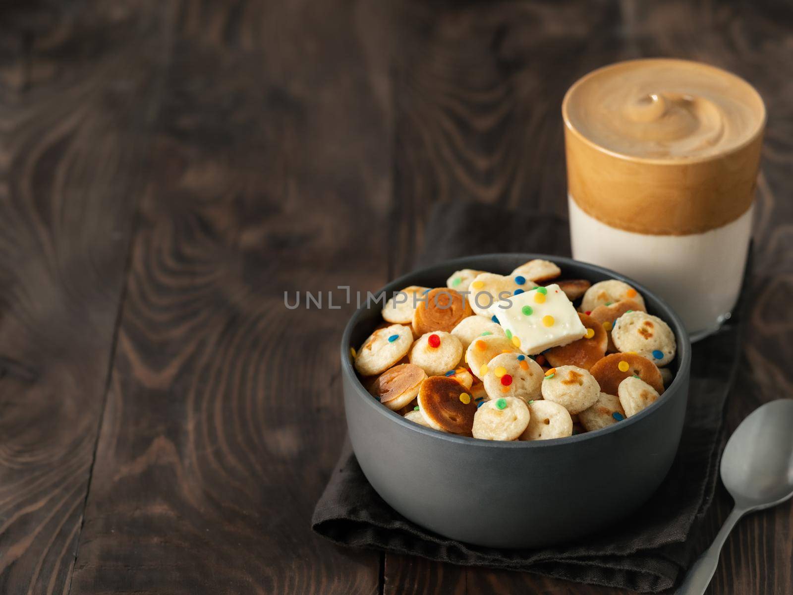 Mini pancakes cereal and dalgona coffee on brown wooden background, copy space. Trendy food and drink - tiny panckakes served sprinkles and whipped instant coffee with milk or korean dalgona coffee
