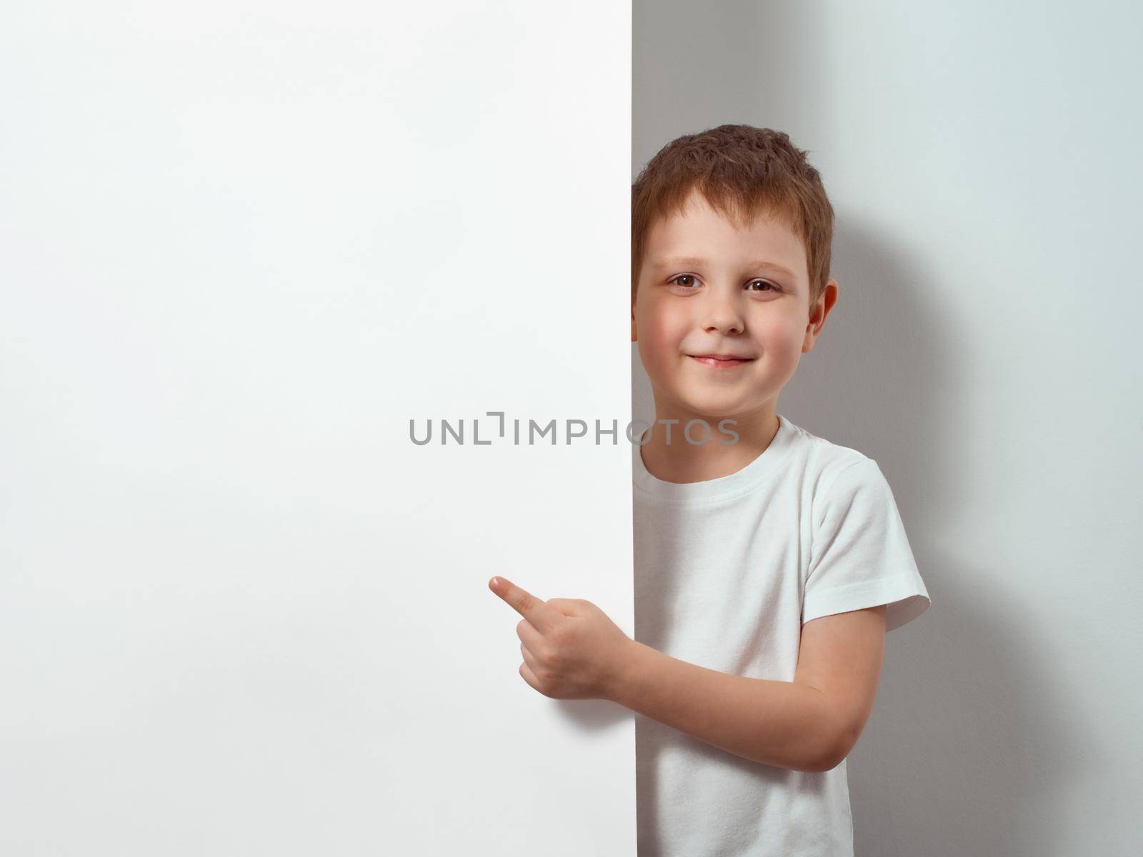 Four-year-old boy shows finger on blank white sheet. Happy child in white t-shirt over white background with copy space for message, mock up
