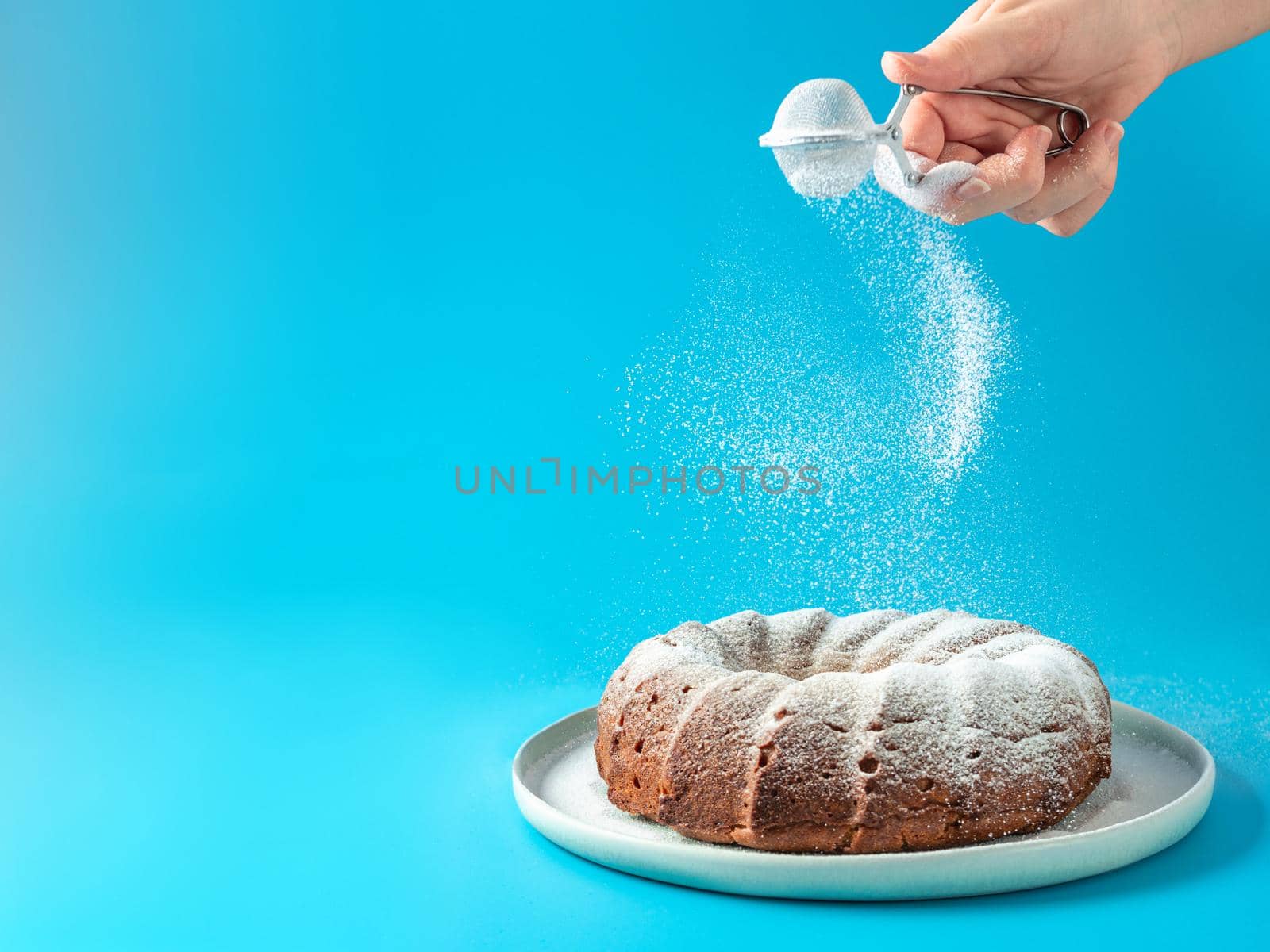 Woman's hand sprinkling icing sugar over fresh home made bundt cake. Powder sugar falls on fresh perfect bunt cake over blue background. Copy space for text. Ideas and recipes for breakfast or dessert