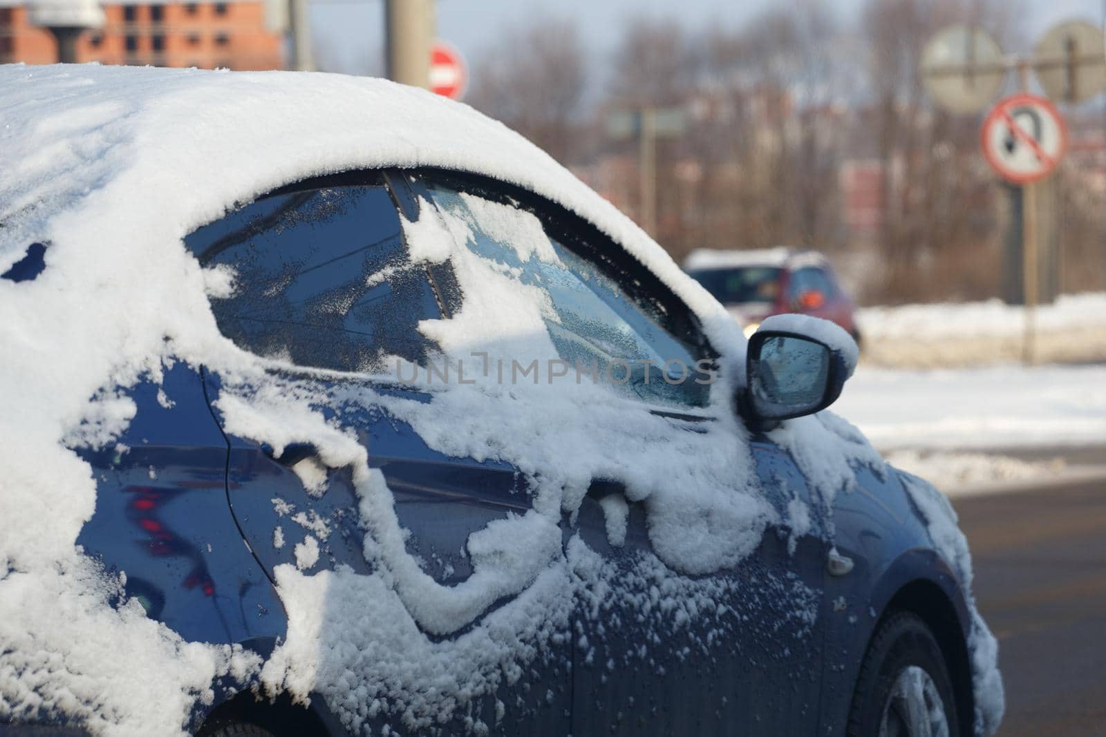 blue car covered with snow riding on winter day street - partially cleaned, selective focus with background blur