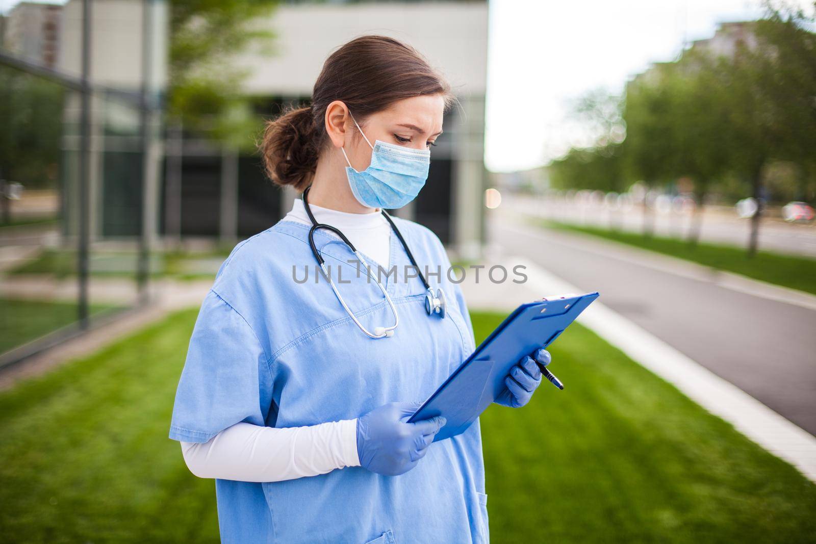 Female UK NHS doctor holding blue clipboard standing outside hospital clinic street entrance by Plyushkin
