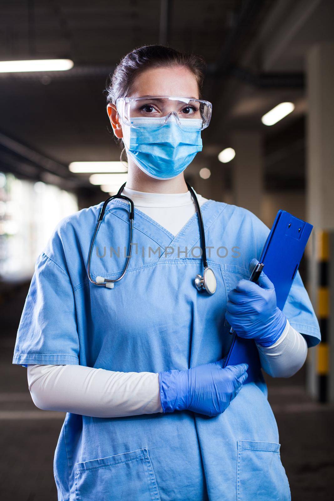 Portrait of young pretty female doctor wearing blue uniform,safety goggles & face mask,worried,overworked & stressed,UK NHS ICU medical key frontline worker fighting COVID-19 pandemic outbreak crisis