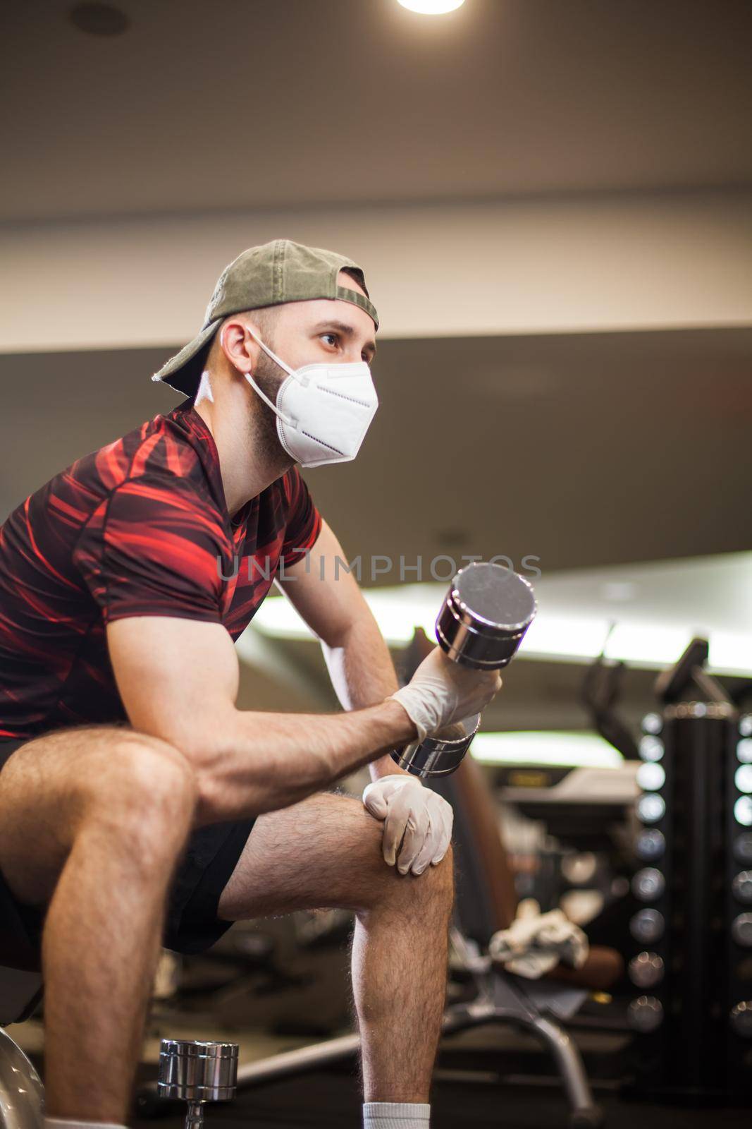 Young caucasian man working out wearing face mask & latex rubber gloves,performing bicep curl with dumbbells,COVID-19 pandemic social distancing rules while working out in indoor gym,United States US