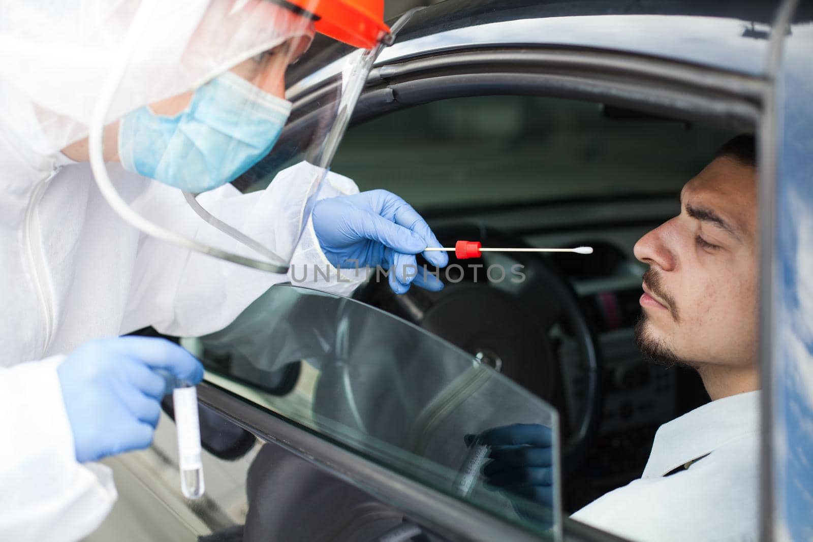 Drive-thru Coronavirus COVID-19 test,paramedic in personal protective equipment & face shield performing nasal swab through car window on male patient sitting in vehicle,mobile US testing site center