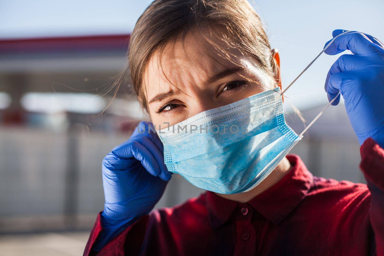 Worried young woman putting PPE face mask on/taking it off in public,wearing protective blue latex surgical gloves,looking at camera with stress and anxiety,new normal for everyday life after COVID-19