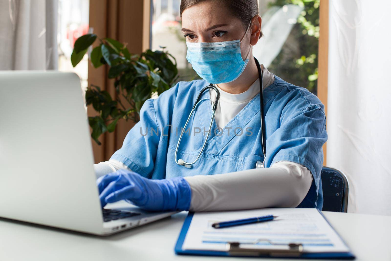 Young caucasian female doctor working on laptop computer in office, wearing protective glove and face mask by Plyushkin