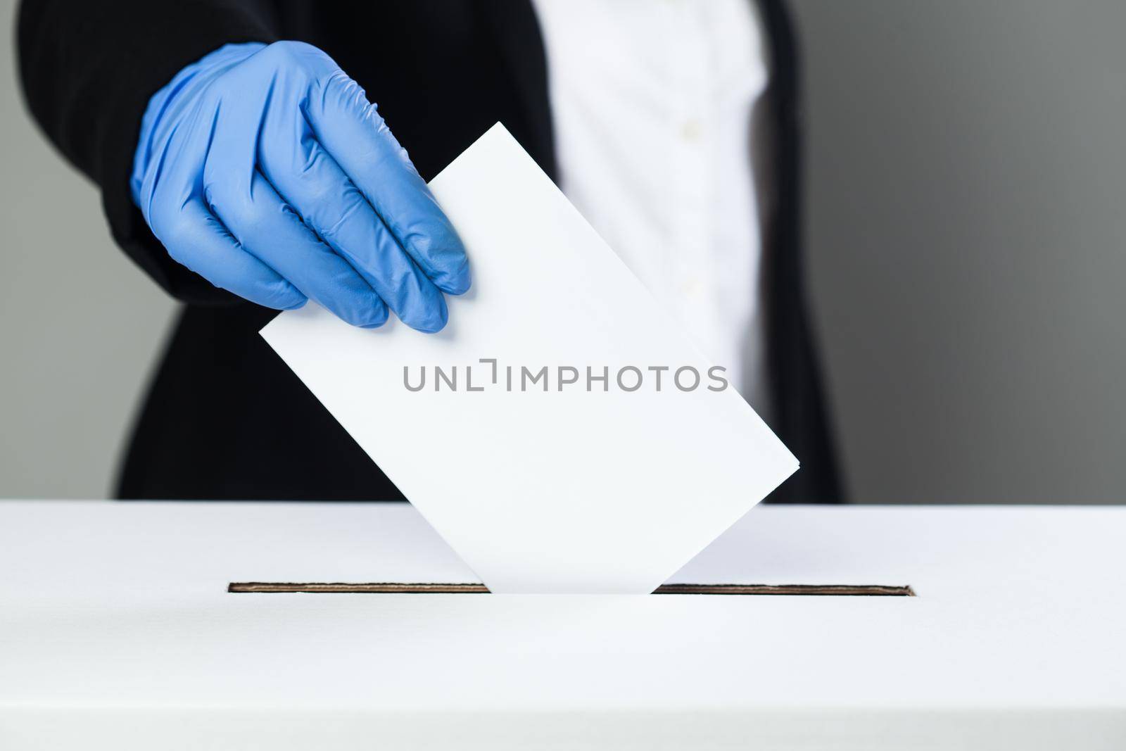 Person in black suit wearing blue protective latex rubber gloves putting ballot paper in vote box, election day during Coronavirus COVID-19 pandemic crisis, prevention and protection at voting place