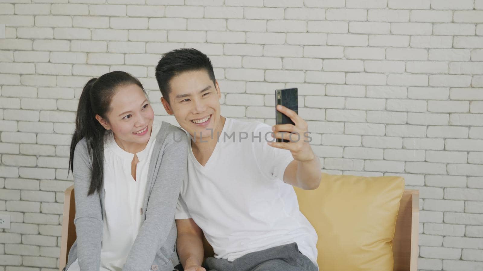 Happy Asian beautiful family couple husband and wife laughing sitting on sofa in the living room taking selfie picture with a smartphone. boyfriend and girlfriend relaxing at home