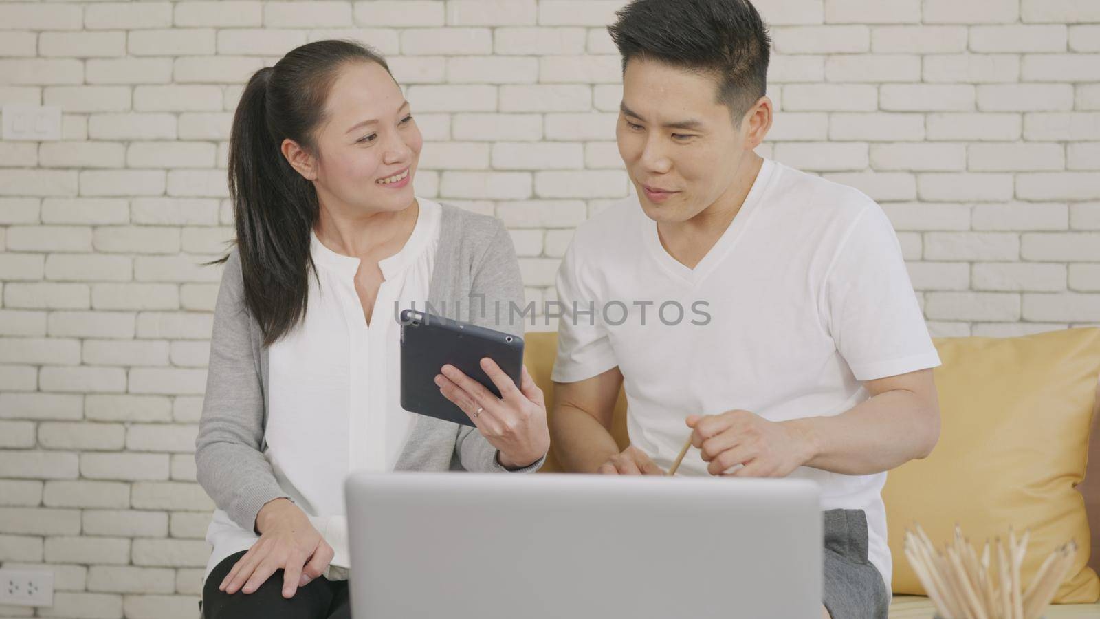 Happy Asian family couple husband and wife work and using digital tablet for discuss. Cheerful loving couple smile shopping something online and watching social media together from home in living room