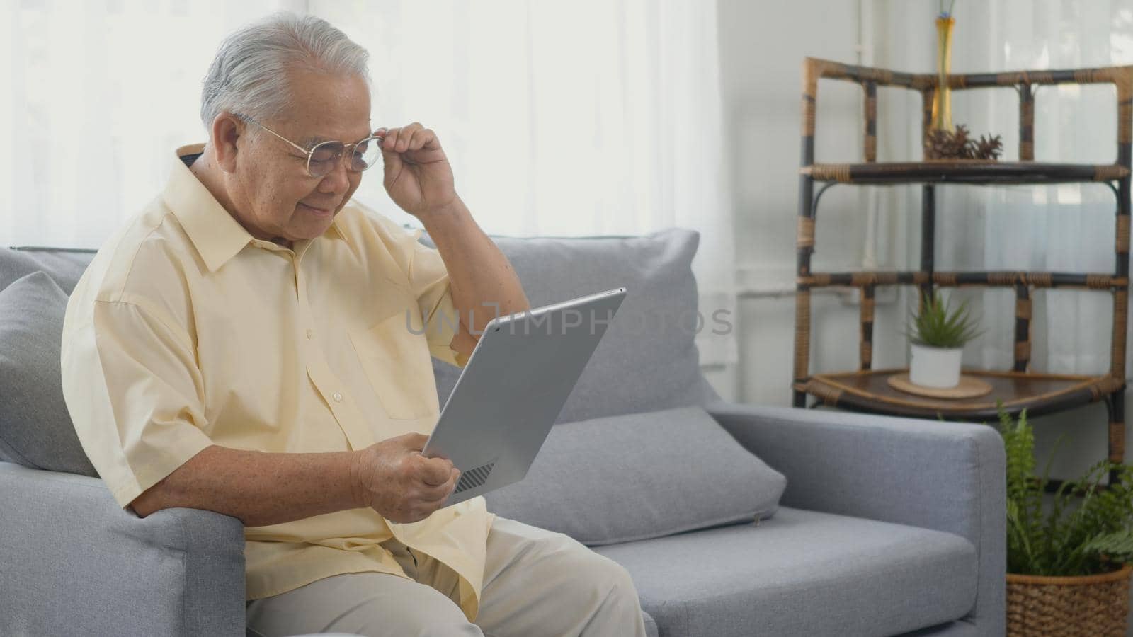 Senior man dressed to wear eyeglasses sitting on sofa looking news on digital tablet by Sorapop