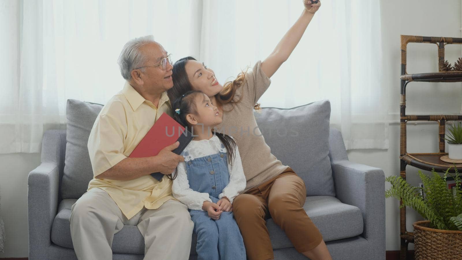 Asian grandparents laughing taking selfie with granddaughter on sofa at home by mobile by Sorapop