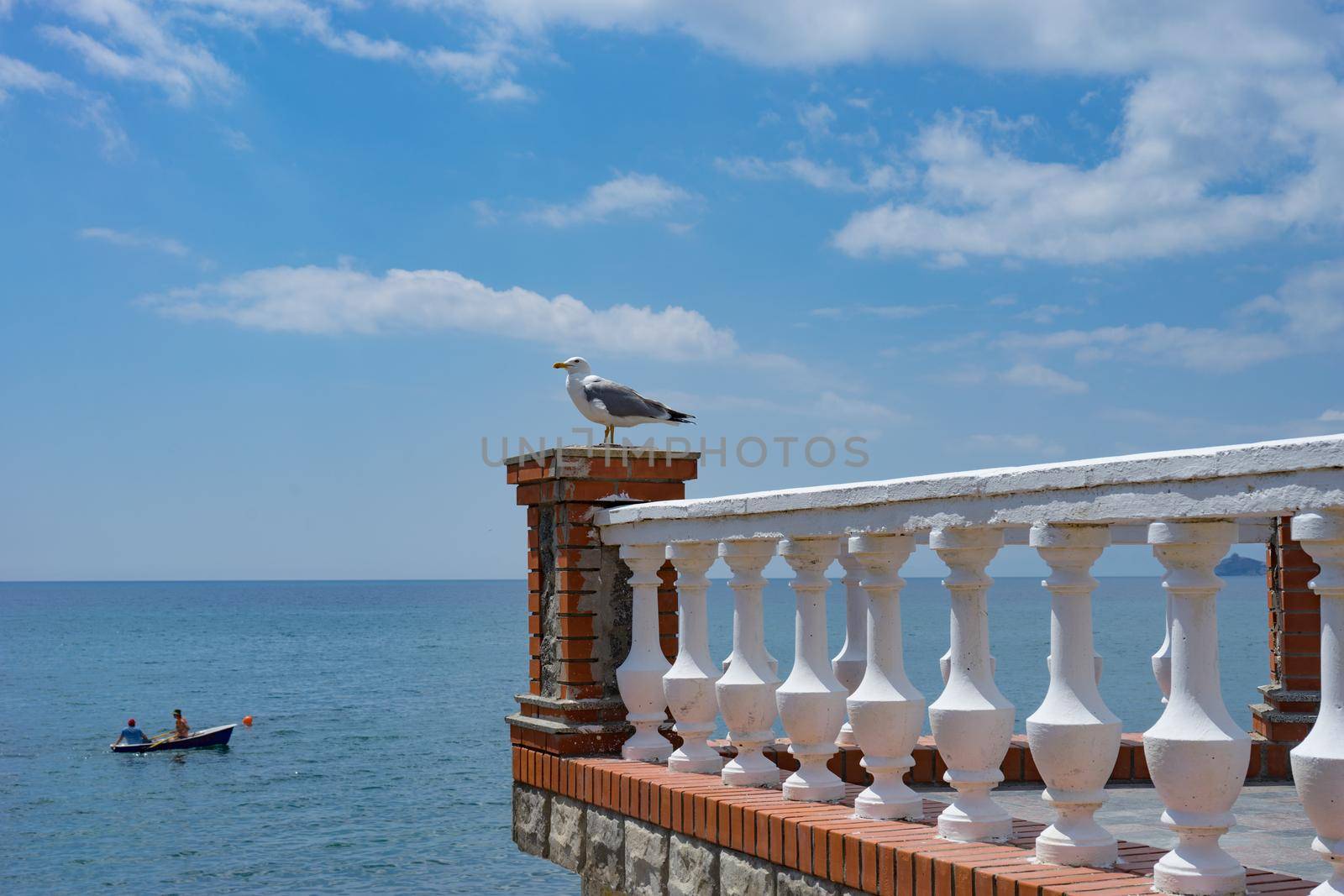 A large Seagull sits on a stone balcony against a blue sea and a boat .